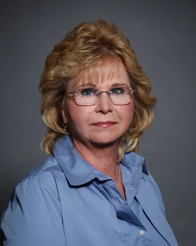 Portrait of a middle-aged woman with blonde hair, glasses, and a blue collared shirt against a gray background.