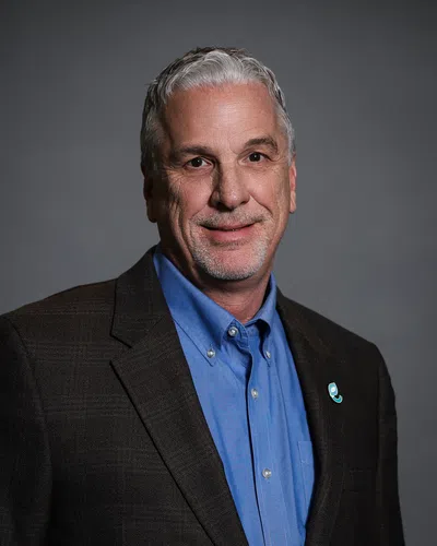 Smiling mature man with gray hair in blue shirt and dark blazer against a plain gray background.