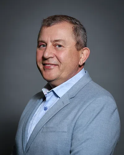 Middle-aged man in light blue blazer and patterned shirt smiling against dark gray background