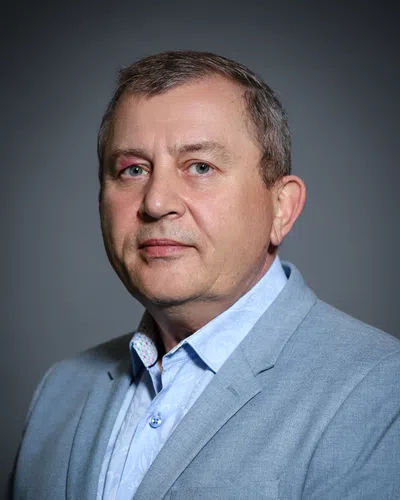 Middle-aged man in a light blue patterned shirt and gray blazer against a dark gray background.