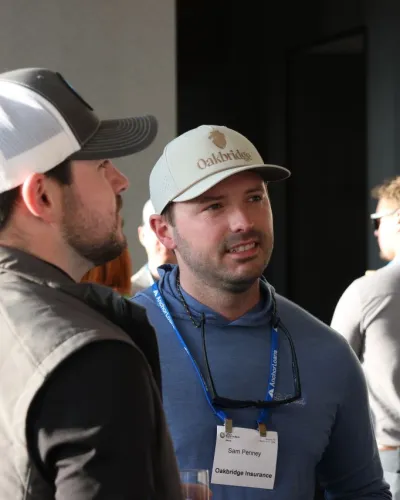 Two men wearing hats and name badges engaged in conversation at a professional event indoors.