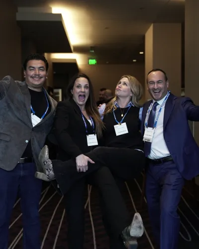 Four professionals at a conference hall posing playfully, smiling and wearing business attire with name badges.