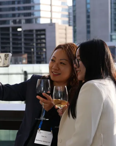 Two women in business attire taking a selfie on a rooftop with wine glasses and city buildings in background