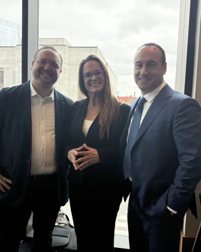 Three professionals in business attire posing and smiling indoors near a window with a museum visible outside.