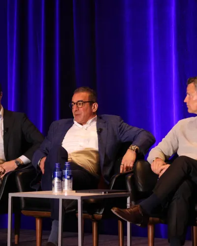 Three men in suits and business casual attire seated on stage chairs during a panel discussion with blue curtains behind.