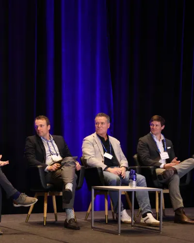 Five male panelists sitting on stage in front of blue curtains during a conference discussion.