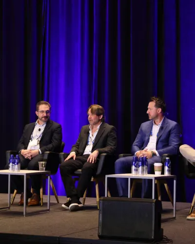 Five men in business attire seated on stage during a panel discussion with purple curtains in the background.