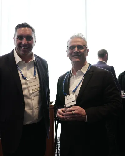 Two men in business attire with name badges at a networking event in a well-lit room with other attendees.