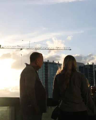 Three people standing on a balcony at sunset with a city skyline and construction crane in the background.