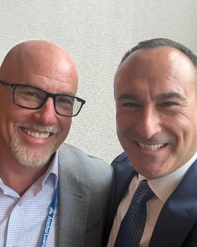Two smiling men in business attire, one wearing glasses and an Anchor Loans badge, posing for a selfie indoors.