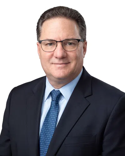 Professional middle-aged man wearing glasses, blue shirt, and dark suit with a blue patterned tie, white background