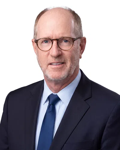 Middle-aged man with glasses wearing a suit, white shirt, and patterned blue tie, smiling slightly against white background.