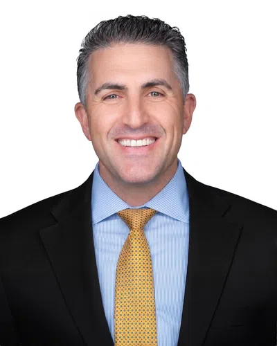 Smiling middle-aged man with gray hair wearing a black suit, blue striped shirt, and yellow patterned tie against white background
