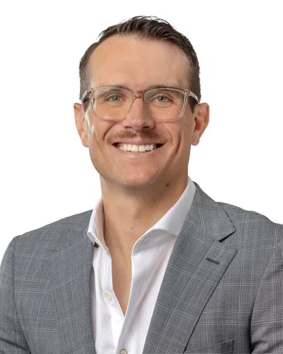 Smiling man wearing clear glasses, a gray plaid blazer, and a white shirt against a white background