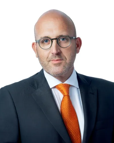 Bald man with glasses wearing a dark suit and bright orange tie against a white background