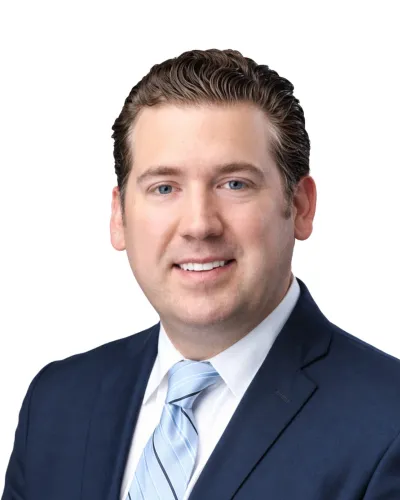 Professional man with brown hair wearing navy suit and light blue striped tie smiles against white background