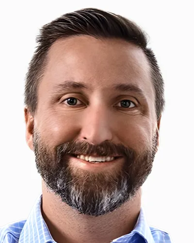 Smiling man with dark hair and beard wearing a light blue checkered shirt on white background