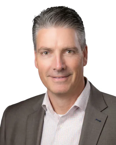Professional headshot of a middle-aged man in a gray suit jacket and checkered shirt smiling against white background
