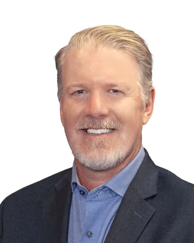 Smiling man with short blond hair wearing a dark suit, light blue shirt, and patterned blue tie against white background