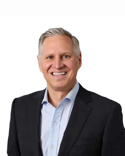 Close-up of a smiling man with light hair, blue eyes, wearing a gray blazer and white shirt against a white background.