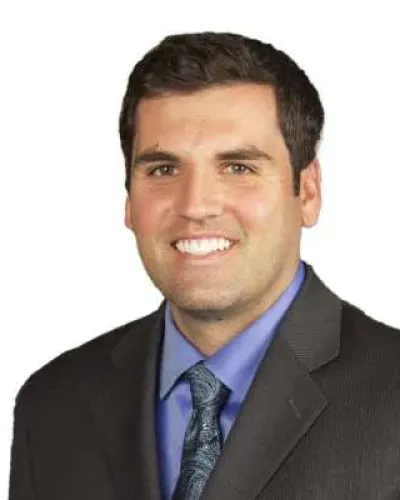 Professional man smiling in a dark suit, blue shirt, and patterned tie against a white background