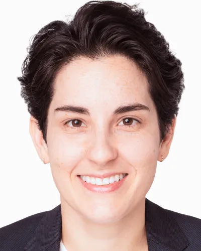 Smiling person with short dark hair wearing a dark blazer against a white background.