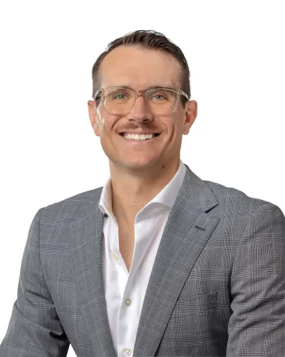 Smiling man wearing clear glasses, a gray plaid blazer, and a white shirt against a white background