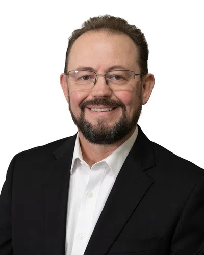Smiling middle-aged man with glasses and beard wearing a black suit jacket and white shirt on a white background