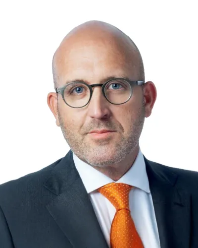 Bald man with glasses wearing a dark suit and bright orange tie against a white background