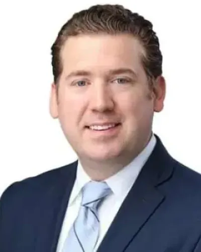Professional headshot of a man in a dark suit, light blue shirt, and tie smiling against a white background