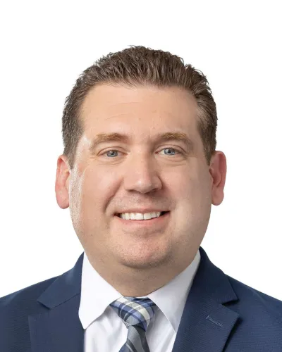Professional headshot of a man in a dark suit, light blue shirt, and tie smiling against a white background