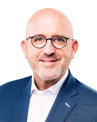 Bald man with glasses wearing a dark suit and bright orange tie against a white background
