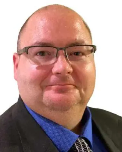 Middle-aged man wearing glasses, blue shirt, patterned tie, and black suit jacket smiling against white background