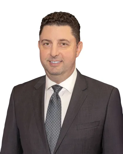 Man with curly hair wearing a dark gray suit, white shirt, and patterned tie smiling against white background