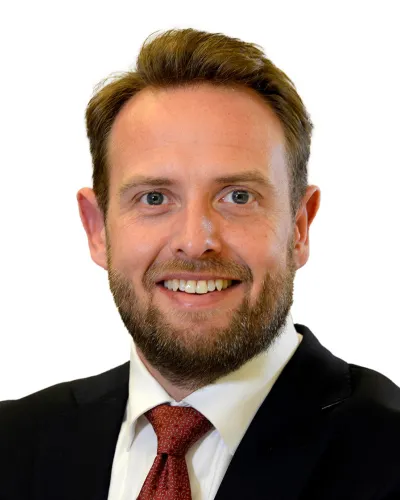 Smiling man with beard wearing a black suit, white shirt, and red tie on a white background.