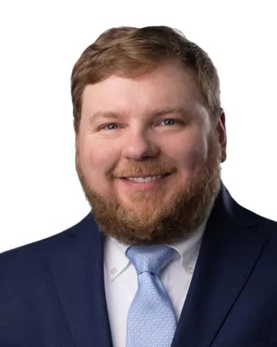 Smiling man with red beard wearing a dark blue suit and light blue tie on a white background