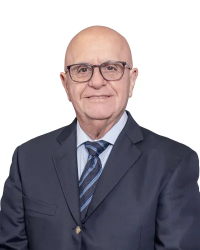 Smiling bald elderly man wearing glasses, a dark suit jacket, light blue shirt, and striped tie on white background