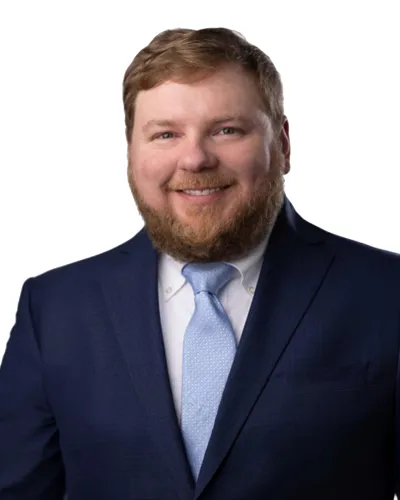 Smiling man with red beard wearing a dark blue suit and light blue tie on a white background