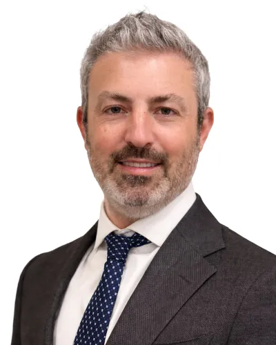 Smiling mature man with gray hair and beard wearing dark suit, white shirt, and blue patterned tie against white background