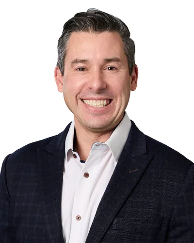 Close-up of a smiling man with short dark hair wearing a white shirt and dark textured jacket against white background