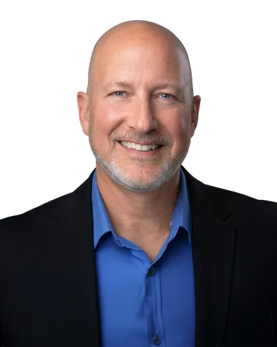 Smiling bald man with light facial hair wearing a blue shirt and dark jacket on a white background.