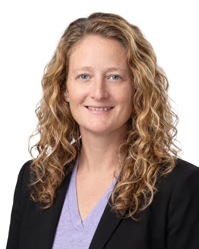 Smiling woman with curly blonde hair wearing a black blazer and lavender V-neck shirt against white background