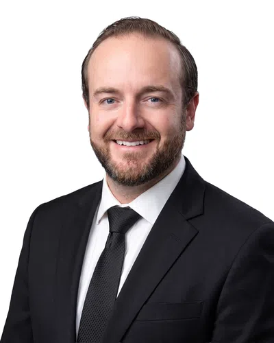 Smiling man with beard wearing black suit, white shirt, and black tie against a white background.