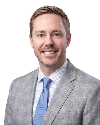 Smiling man with light brown hair wearing a gray checkered suit and blue tie on white background