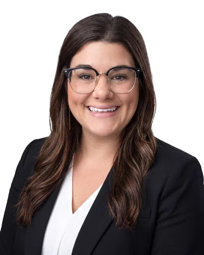 Smiling woman with long brown hair, wearing glasses, black blazer, and white blouse on white background