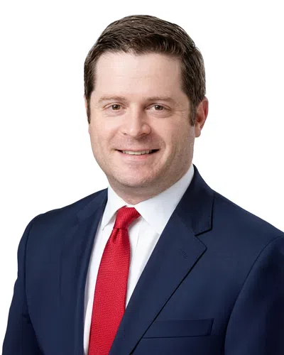 Smiling man in a navy blue suit, white shirt, and red tie against a white background.