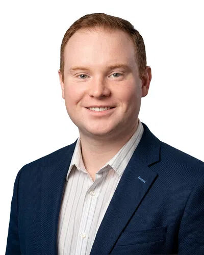Headshot of a smiling man with red hair wearing a blue blazer and striped shirt on a white background