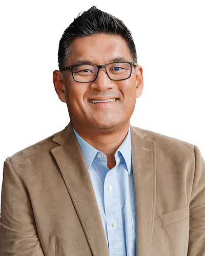 Smiling man wearing glasses, light blue shirt, and brown blazer against a white background