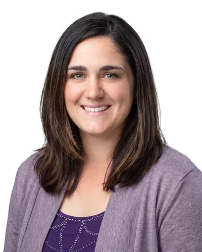 Smiling woman with shoulder-length dark hair wearing a purple top and cardigan against a white background