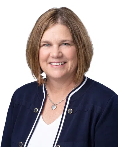 Smiling middle-aged woman with short brown hair wearing a navy blazer and heart pendant necklace against white background